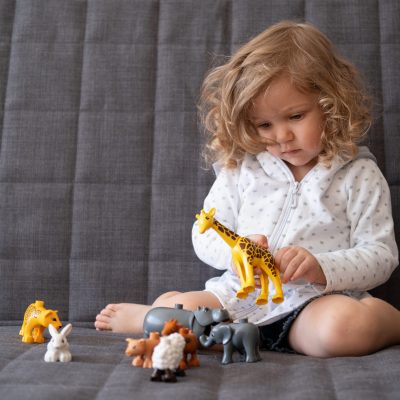 Cute curly toddler girl playing with with toy zoo sitting on couch. Toys for little kids. Child with educational toy. Early development